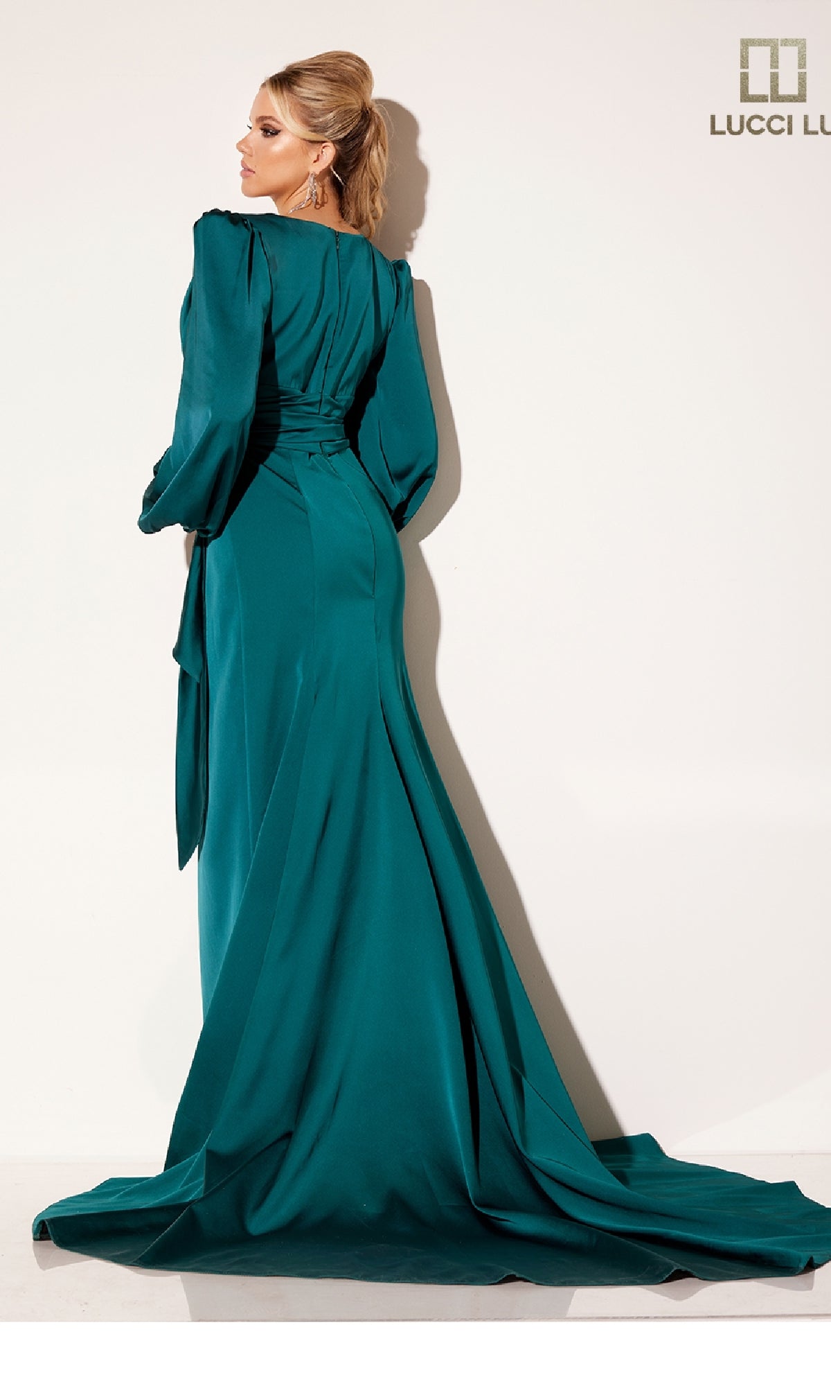 A woman stands facing a white wall in the Lucci Lu C8088 long formal dress, a teal, backless gown with long sleeves, a wrap-style skirt, and a flowing train.